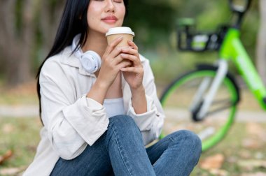 Parktaki çimlerin üzerinde dizlerinin üzerinde otururken kahve kokusu alan Asyalı bir kadını yakından çek. Doğada dinlenme, açık hava aktivitesi, yeşil orman parkı..