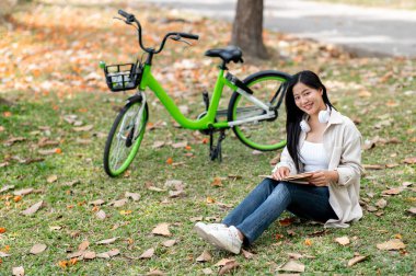 Asyalı bir kadın, kitabı kucağında okurken gülümsüyor, çim tarlasında oturuyor. Açık hava aktivitesi, Yeşillik Orman Parkı.