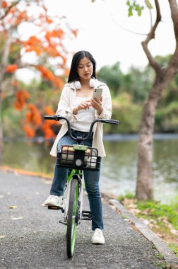 Parktaki göl yolu üzerinde bisiklet süren Asyalı bir kadın telefonuna bakmak için bisikleti durdurdu. Açık hava aktivitesi, Yeşillik Orman Parkı.