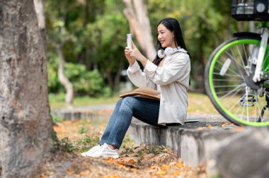 Gülümseyen Asyalı bir kadın park kaldırımında otururken telefonuyla fotoğraf çekiyor. Selfie ya da sahne fotoğrafı çekmek, açık havada takılmak, yeşil orman parkı..