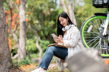 Gülümseyen Asyalı bir kadın park kaldırımında otururken telefonunda daktilo yazıyor. Sohbet etmek ya da fotoğraf göndermek, dışarıdaki yeşil orman parkında takılmak..