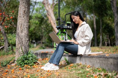 Asyalı bir kadın dizüstü bilgisayarı kucağında kullanıyor, parkta ya da bahçede çalışıyor ya da ders çalışıyor. Güzel Yeşillik.