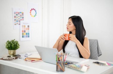 Asyalı bir kadın iki eliyle bir kupayı tutarken duvardaki renk çarkına ya da renk paletine bakıyor. Ofiste çalışan İç mimar veya Grafik Tasarımcı.