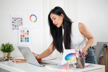 Bir stil ve basılı renk tablosu tutarken dizüstü bilgisayara bakmak için masaya eğilen Asyalı bir kadın. İç Tasarımcı veya Grafik Tasarımcısı.
