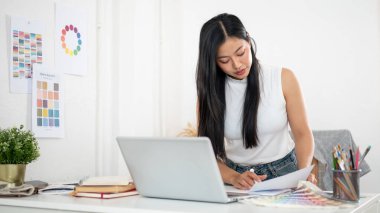 Asyalı bir kadın diğer çalışma materyalleri arasında yer alan belgeleri okumak için masanın üzerine eğiliyor. İç Tasarımcı veya Grafik Tasarımcısı.