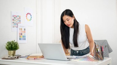 Asyalı bir kadın dizüstü bilgisayar ekranına daha yakından bakmak için masanın üzerine eğiliyor. İç Tasarımcı veya Grafik Tasarımcısı.