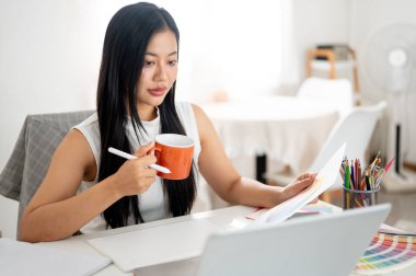 Asyalı bir kadın dizüstü bilgisayar ekranına bakarken elinde turuncu bir seramik bardak tutuyor. İç Tasarımcı veya Grafik Tasarımcısı.