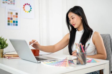 Asyalı bir kadın masadaki dizüstü bilgisayara bakıyor ve bir stilus kalem doğrultuyor. Ofiste çalışan İç mimar veya Grafik Tasarımcı.