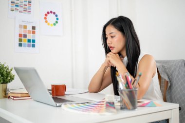 Asyalı bir kadın iki elini de çenesinin altına koyuyor ve masadaki dizüstü bilgisayara dikkatle bakıyor. Ofiste çalışan İç mimar veya Grafik Tasarımcı.