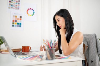 Asyalı bir kadın, grafik tabletle tasarım üzerinde çalışırken dikkatle dizüstü bilgisayara bakıyor. Ofiste çalışan İç mimar veya Grafik Tasarımcı.