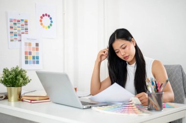 Asyalı bir kadın çalışma masasındaki tasarım rengiyle ilgili belgeleri sakince okuyor. Ofiste çalışan İç mimar veya Grafik Tasarımcı.