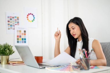 Asyalı kadın dizayn renkleriyle ilgili belgeleri tutarken dizüstü bilgisayara bakıyor. Ofiste çalışan İç mimar veya Grafik Tasarımcı.