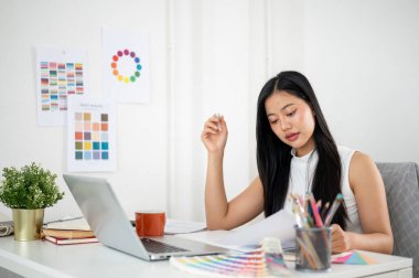 Asyalı bir kadın, grafik tablet ve dizüstü bilgisayar üzerinde tasarım yaparken belgeleri okuyor. İç Tasarımcı veya Grafik Tasarımcısı ofiste.