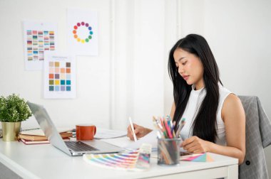 Asyalı kadın dokümanları okurken grafik tablet üzerinde çizim yapıyor ya da tasarım yapıyor. İç Tasarımcı veya Grafik Tasarımcısı ofiste.