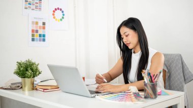 Asyalı kadın grafik tablet üzerinde çizim yapıyor ya da tasarım yapıyor ve dizüstü bilgisayarın klavyesindeki tuşa basıyor. İç Tasarımcı veya Grafik Tasarımcısı.