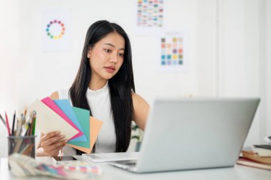 Asyalı kadın dizüstü bilgisayarda kağıt rengi çizelgelerinden seçilmiş renkle çalışıyor. İç Tasarımcı veya Grafik Tasarımcısı.