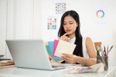 Asyalı kadın kağıt rengi çizelgelerinden rengini seçerken derin düşünüyor. Çalışma masasında dizüstü bilgisayarla oturuyor. İç Tasarımcı veya Grafik Tasarımcısı.