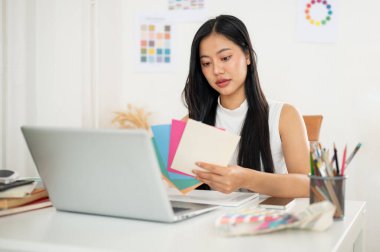 Asyalı bir kadın, dizaynında kullanmak için kağıt rengi çizelgelerinden bir renk seçiyor. Çalışma masasında dizüstü bilgisayarla oturuyor. İç Tasarımcı veya Grafik Tasarımcısı.