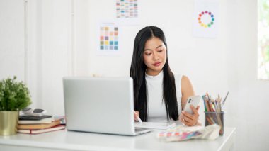 Asyalı bir kadın telefona bakarken bir stilus kalem çiziyor ya da tablete yazı yazıyor. Ofiste çalışan İç mimar veya Grafik Tasarımcı.