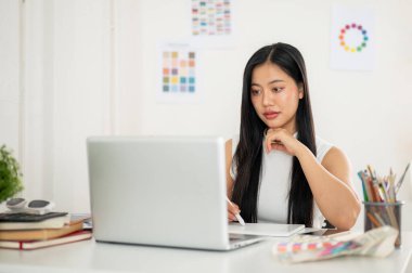 Asyalı bir kadın dizüstü bilgisayar ekranına bakarken bir stilus kalem çiziyor ya da tablete yazıyor. Ofiste çalışan İç mimar veya Grafik Tasarımcı.