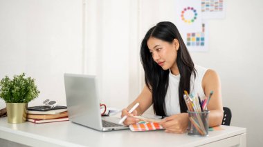 Asyalı bir kadın dizüstü bilgisayara bakarken renk çizelgelerinden ya da paletlerden renk seçiyor. Ofiste çalışan İç mimar veya Grafik Tasarımcı.