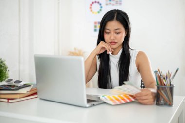 Asyalı bir kadın renk grafiklerine ya da renk paletlerine dikkatlice bakıyor. Ofiste çalışan İç mimar veya Grafik Tasarımcı.