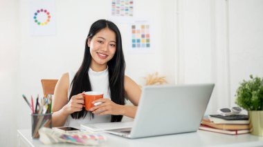 Gülümseyen Asyalı bir kadın iki eliyle bir kupa tutuyor. Üzerinde çalışma materyalleri olan bir dizüstü bilgisayarın önünde oturuyor. İç Tasarımcı veya Grafik Tasarımcısı.