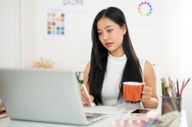 Asyalı bir kadın elinde stilus kalem çizimi ya da tablet üzerinde bir elinde bardak tutan bir yazı tutuyor. Ofiste çalışan İç mimar veya Grafik Tasarımcı.