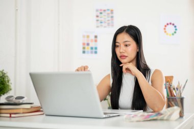 Asyalı bir kadın çalışma materyallerinin olduğu masadaki dizüstü bilgisayara bir stilus kalem doğrultuyor. Ofiste çalışan İç mimar veya Grafik Tasarımcı.