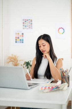 Asyalı bir kadın bir elini çenesinin altına koyarak bir stilus kalem çiziyor ya da tablete yazı yazıyor. Ofiste çalışan İç mimar veya Grafik Tasarımcı.