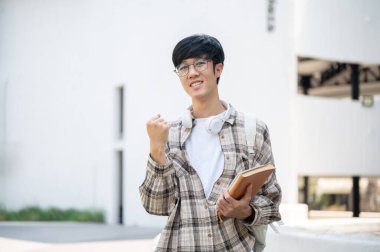 Gözlüklü Asyalı bir adam bir binanın önünde dururken kitap tutuyor ve yumruk atıyor. Mutlu ya da heyecanlı görünüyorsun. Dışarıda Yürüyüş.