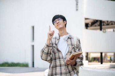 Gözlüklü Asyalı bir adam bir binanın önünde dururken kitapları tutuyor ve parmağını kaldırıyor. Düşünceli olmak mı yoksa bir şeyler bulmak mı?.