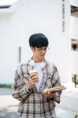 Gözlüklü Asyalı bir adam bir kahve fincanı tutuyor ve binanın önünde dururken kitaplarına bakıyor. Dışarı çıkmak ya da bir yerlere gitmek..