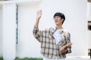 Gözlüklü Asyalı bir adam bir binanın önünde dururken kitapları tutuyor ve el sallıyor. Selamla ya da gönder.