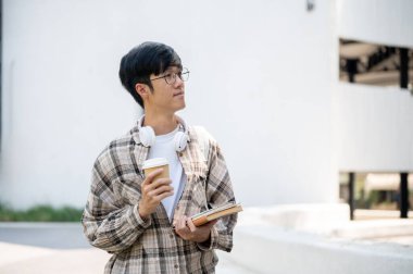 Gözlüklü Asyalı bir adam binanın etrafına bakarken elinde kitaplarla bir kahve fincanı tutuyor. Dışarı çıkmak ya da bir yerlere gitmek..
