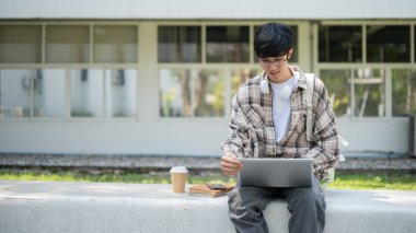 Gözlüklü Asyalı bir adam elinde kahve fincanı ve kitaplarla parkta beton bir bankta otururken dizüstü bilgisayar tutuyor. Çalış ya da dışarıda çalış.