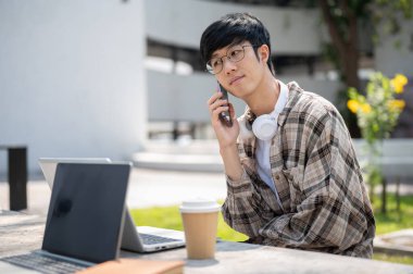 Gözlüklü Asyalı bir adam parktaki beton bir masada dizüstü bilgisayarla otururken akıllı telefonuyla arama yapıyor. Çalış ya da dışarıda çalış.