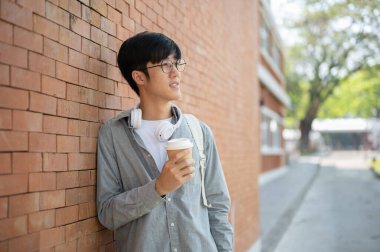 Gözlüklü gülümseyen Asyalı bir adam bir kahve fincanı tutuyor ve tuğla duvara yaslanmış bir şekilde duruyor. Biraz dinlen ya da dışarıda takıl..