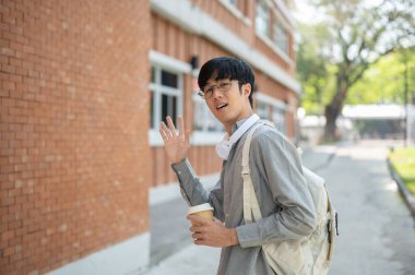 Gözlüklü Asyalı bir adam bir kahve fincanı tutuyor ve tuğla bir duvarın yanında dururken elini sallıyor. Dışarı çıkmak mı yoksa bir yere gitmek mi?.