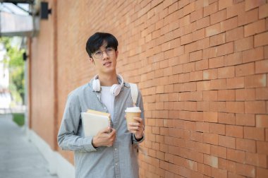 Gözlüklü Asyalı bir adam elinde kitaplarla bir kahve fincanı tutuyor ve tuğla duvarın yanında duran bir kafaya bakıyor. Dışarı çıkmak mı yoksa bir yere gitmek mi?.