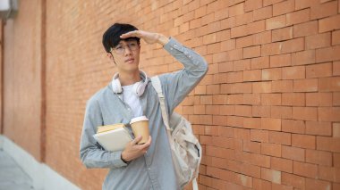 Gözlüklü Asyalı bir adam elinde kitaplarla kahve fincanı tutuyor ve tuğla duvarın yanında dururken gözleri üzerinde duran bir kafaya bakıyor. Dışarı çıkmak mı yoksa bir yere gitmek mi?.