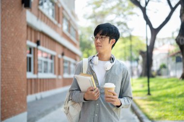 Gözlüklü Asyalı bir adam kahve fincanıyla kitap tutuyor ve tuğla bir duvarın yanında dikilirken kafasını çeviriyor. Dışarı çıkmak mı yoksa bir yere gitmek mi?.