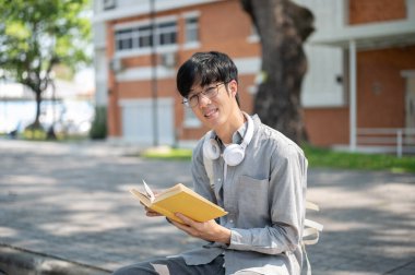Gözlüklü Asyalı bir adam, elinde sarı kapaklı bir kitapla kaldırım bankında otururken gülümsüyor. Boş zaman geçirmek, Serinlemek.