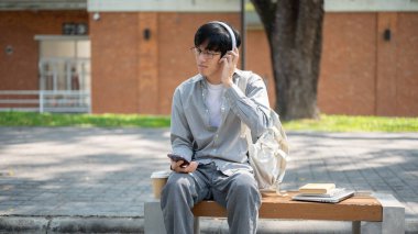 Gözlüklü Asyalı bir adam gözlerini kapatıyor kulaklığıyla müzik dinliyor, kaldırım bankında oturuyor. Boş zaman geçirmek. Dışarısı ürpertici..