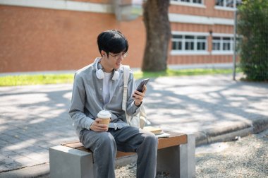 Kaldırımdaki bankta kahve fincanı tutarken telefonuna bakan gülümseyen Asyalı bir adam. Boş zaman geçirmek. Dışarısı ürpertici..