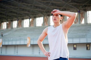 Beyaz kolsuz tişörtlü Asyalı adam bir eli belinde, diğer eli de yarış pistinde koştuktan sonra ter siliyor..
