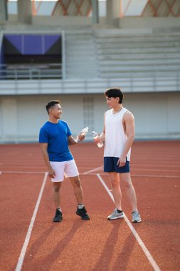 İki Asyalı sporcu su şişelerini tutarken birbirlerine gülümseyip yarış pistinde duruyorlar. Yürüyüş ya da Koşu, Açık hava egzersizi ya da antrenman.
