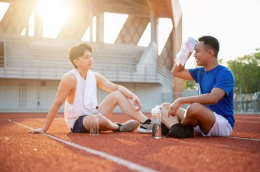 İki Asyalı sporcu dinleniyor, yarış pistinde birlikte oturuyorlar. Koşmak ya da koşmak, dışarıda egzersiz yapmak ya da egzersiz yapmak, spor ve sağlık.