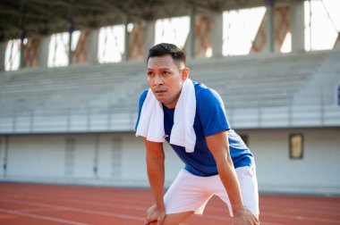 Mavi tişörtlü Asyalı adam elleri dizinde yarış pistinde koştuktan sonra dinleniyor. Açık hava egzersizi ya da eğitim. Spor ve sağlık.