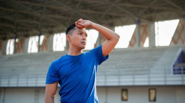 Mavi tişörtlü Asyalı adam ayakta ve terini eliyle siliyor, koştuktan sonra dinleniyor. Açık hava egzersizi ya da eğitim. Spor ve sağlık.